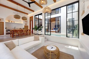 A modern living room with a white sofa, a round wicker coffee table, and large windows offering a view of the outdoors. - Villa Kala (Canggu)