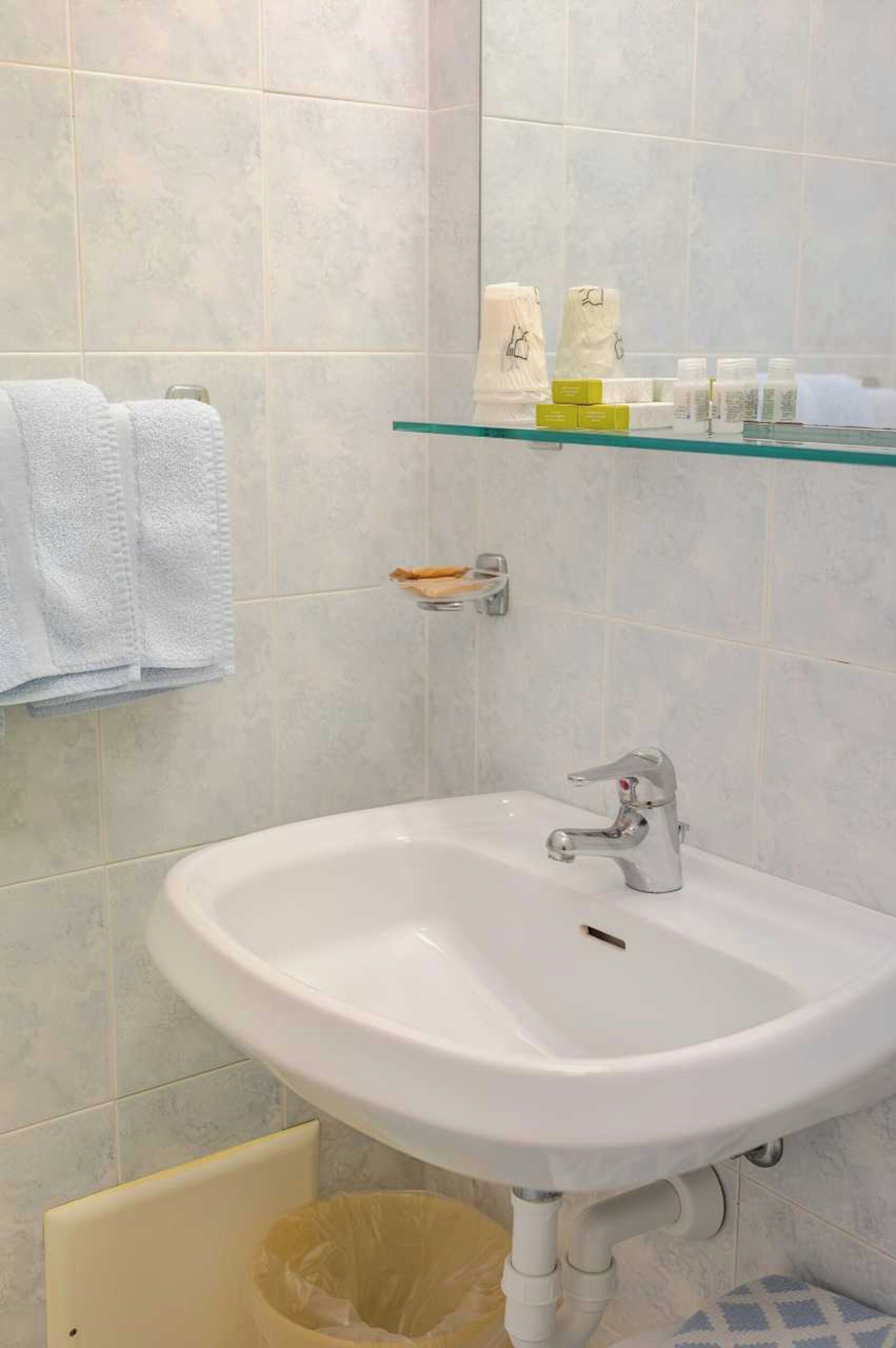 A bathroom with a white sink, a glass shelf above it, and a trash bin below.