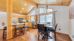 A modern kitchen with wooden cabinets, a central island with bar stools, a dining table with chairs, and a large window with a view of trees.