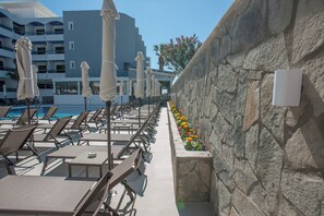 Ein Poolbereich mit Liegestühlen und Sonnenschirmen, eine Steinmauer mit Blumen und ein modernes Gebäude im Hintergrund.