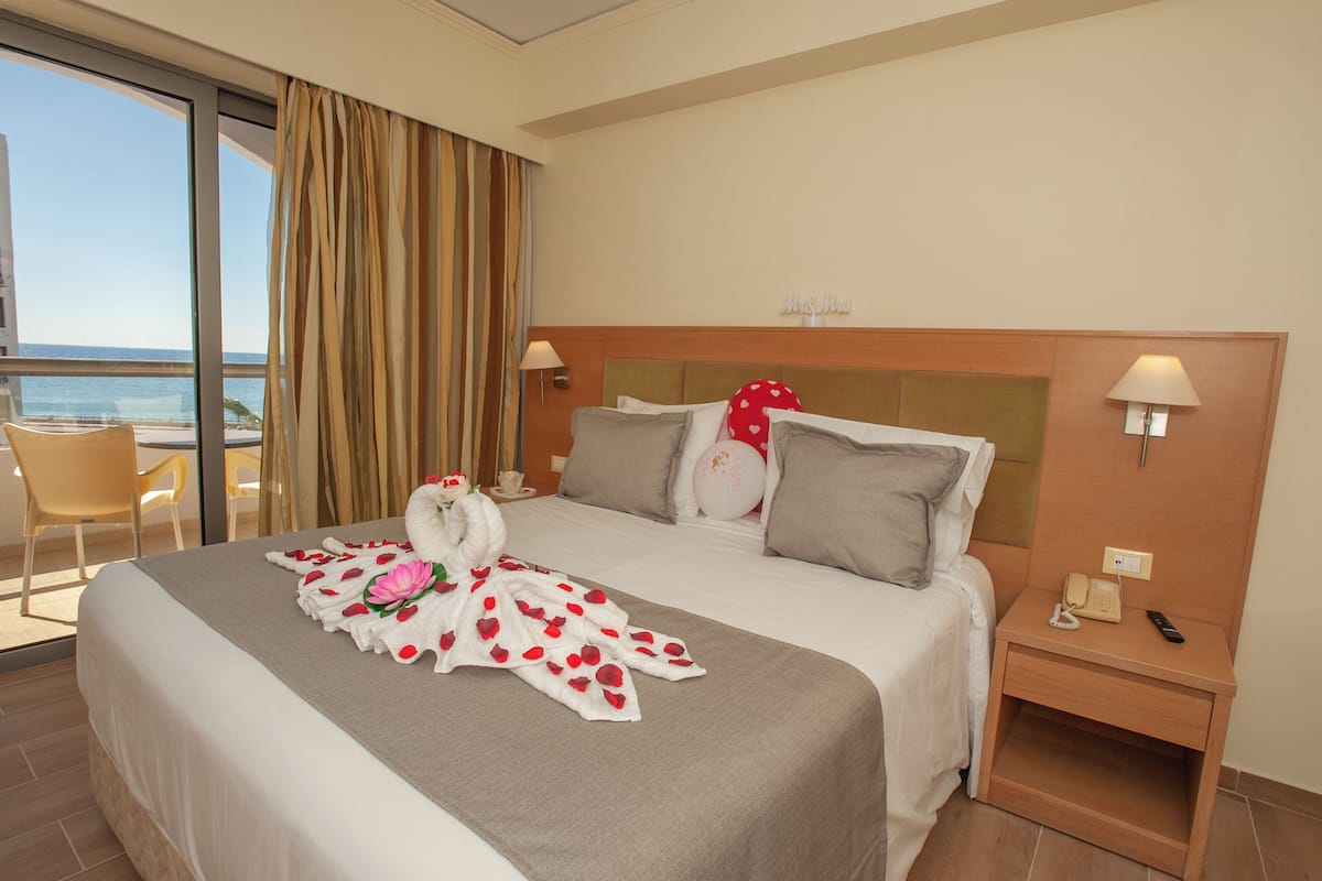 A hotel room with a large bed, a nightstand, a phone, and a view of the sea through a large window.