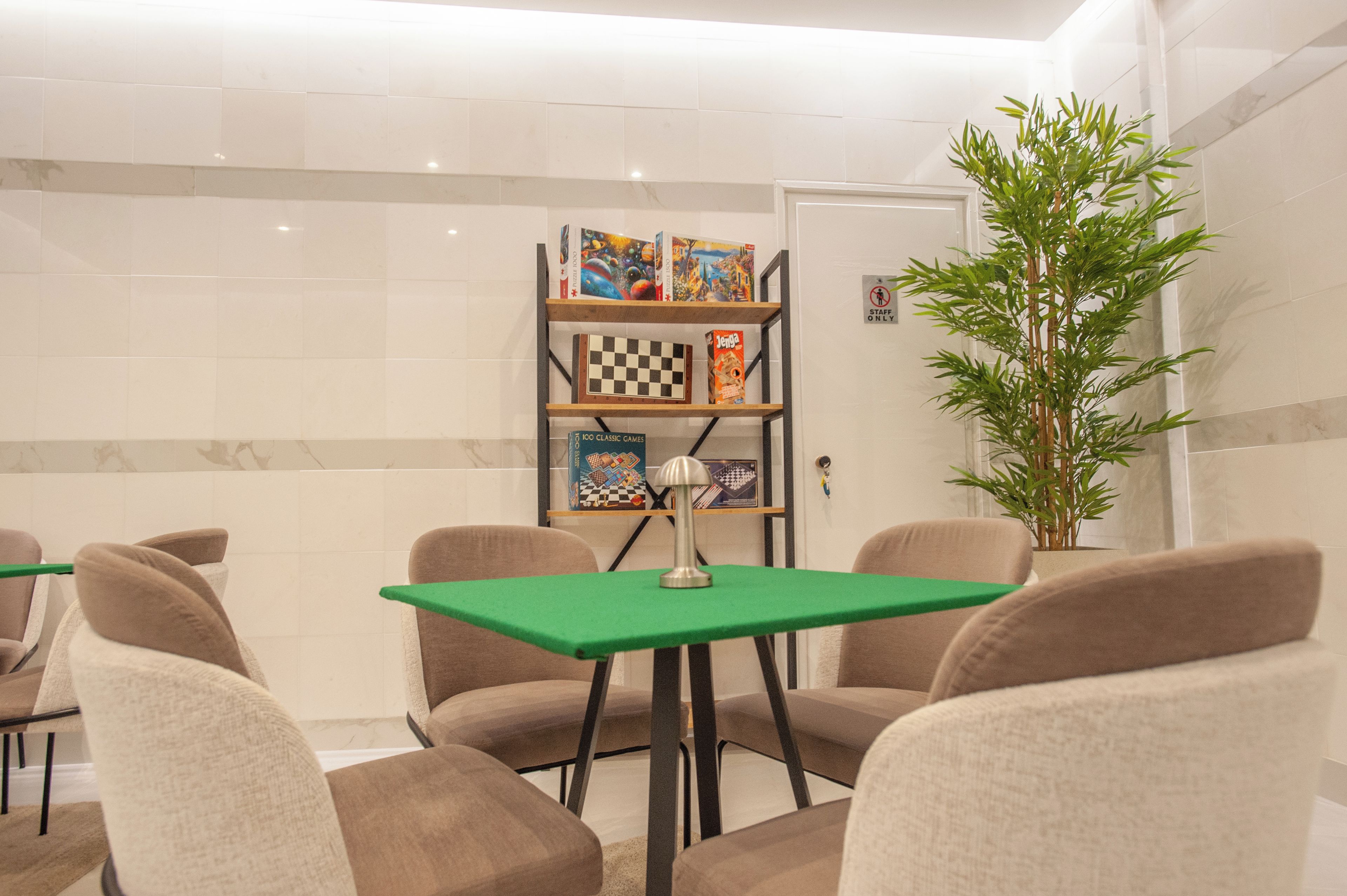 A room with a green table, beige chairs, a bookshelf with board games, and a potted plant.
