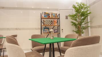 A room with a green table, beige chairs, a bookshelf with board games, and a potted plant.