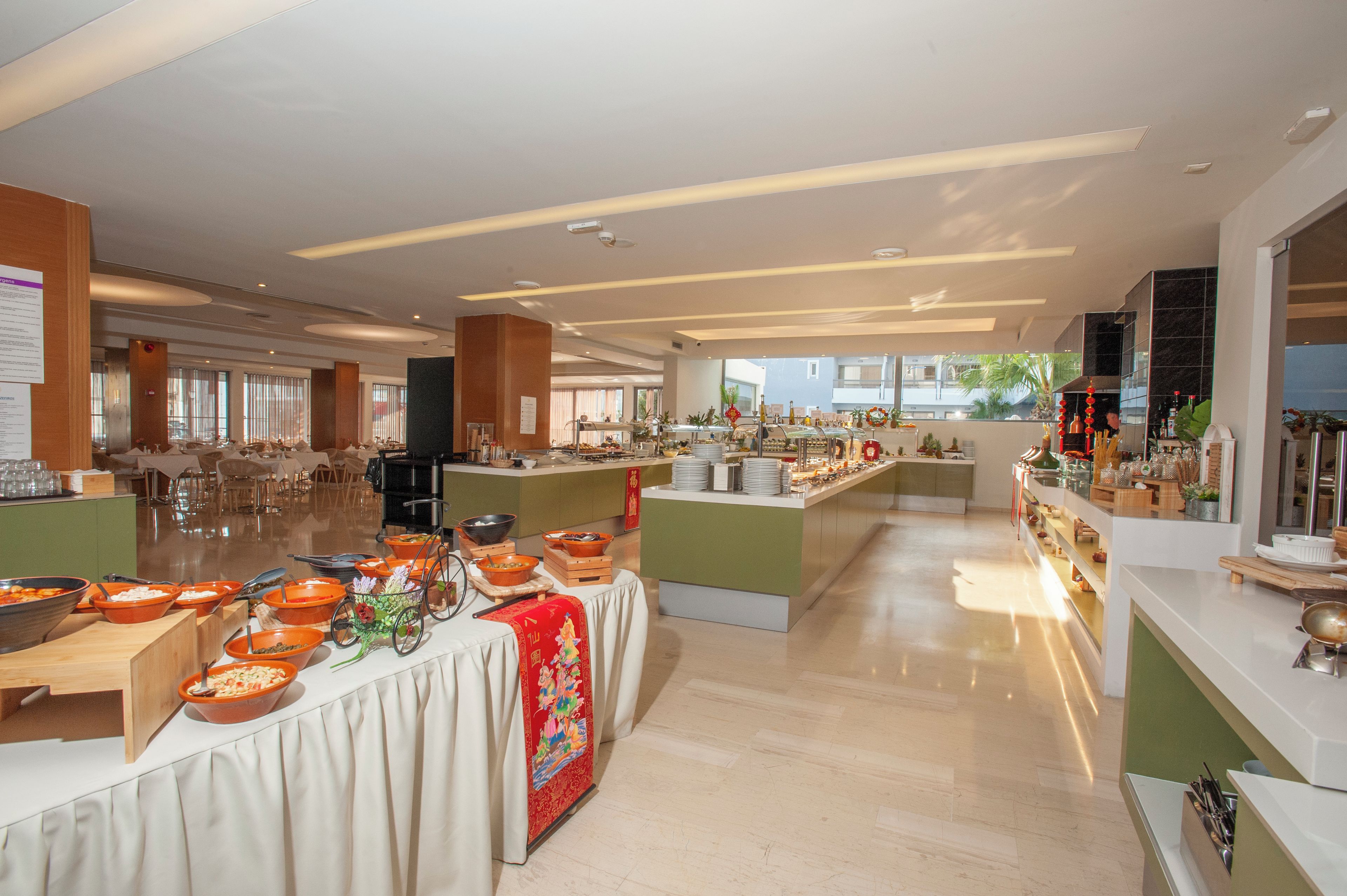 A spacious hotel dining area with multiple buffet stations, a central serving counter, and a view of the outdoors through large windows.