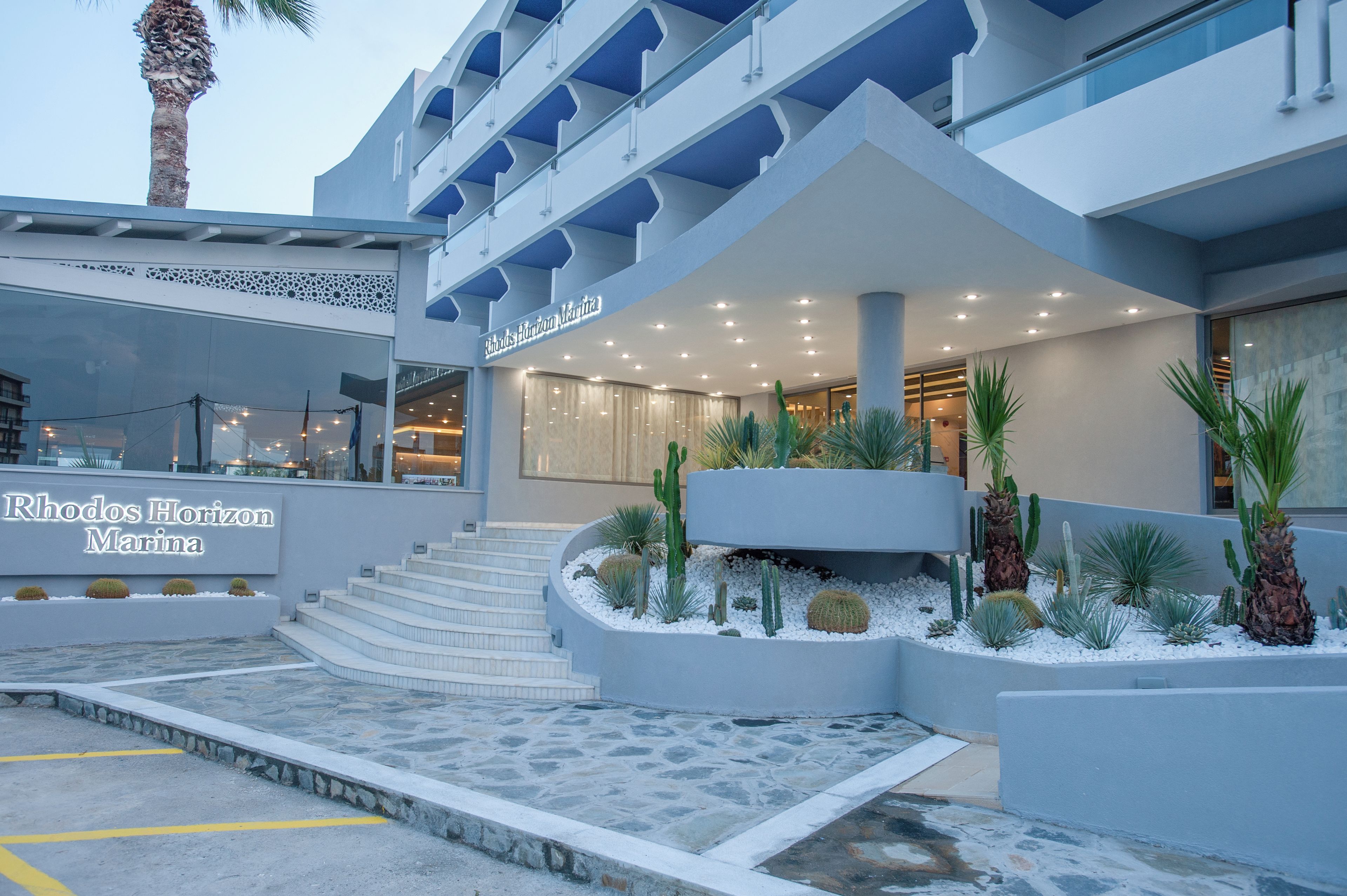 A hotel entrance with a modern design, featuring a staircase leading to the entrance, a sign for 'Rhodos Horizon Marina', and a landscaped area with cacti and palm plants.