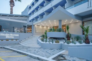 A hotel entrance with a modern design, featuring a staircase leading to the entrance, a sign for 'Rhodos Horizon Marina', and a landscaped area with cacti and palm plants.