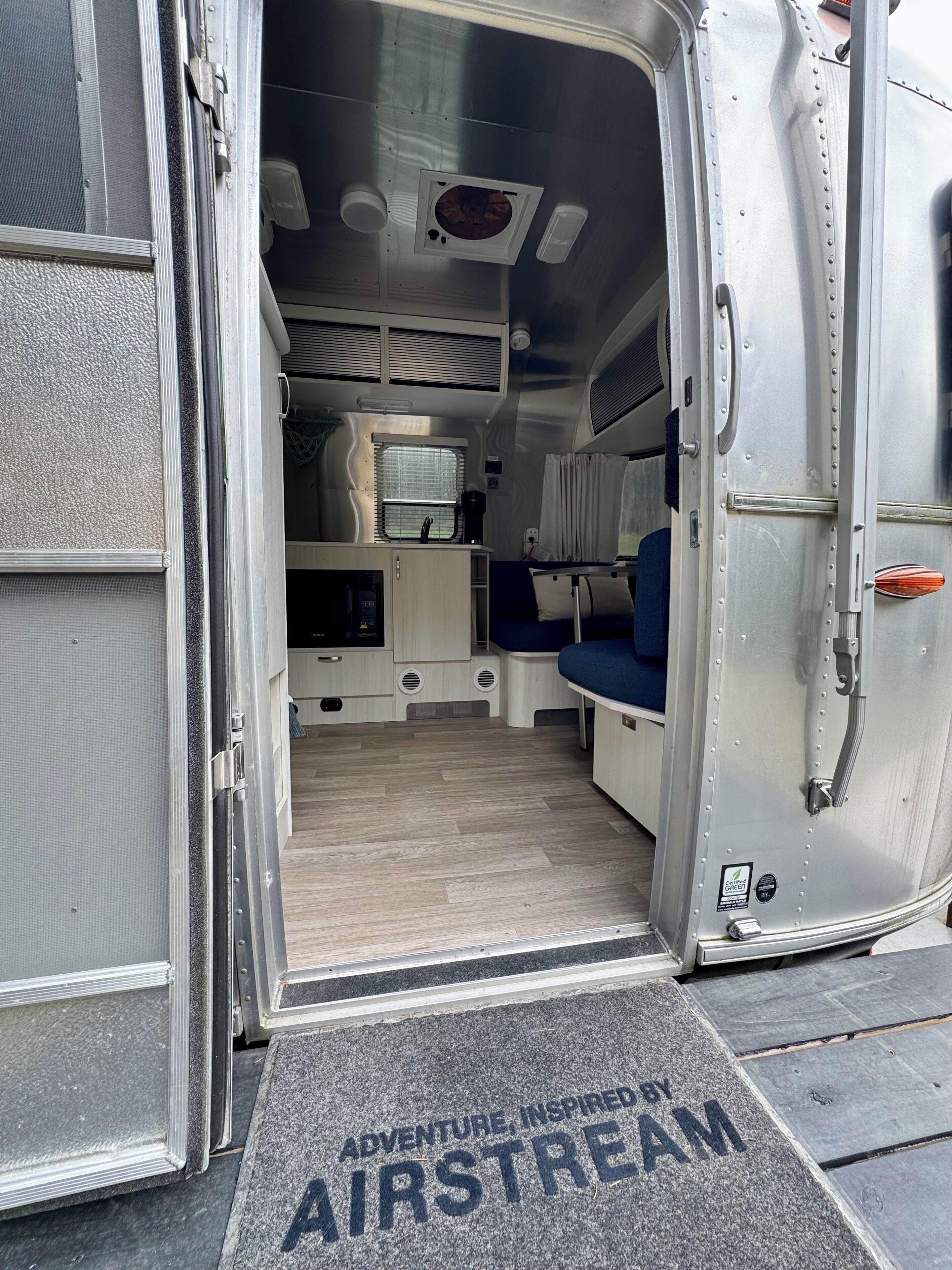 A van interior with a kitchenette, seating area, and a door with a doormat that reads "Adventure Inspired by Airstream."