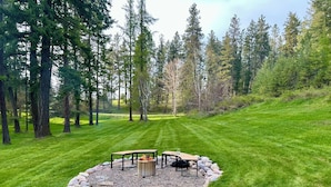 A circular outdoor seating area with a fire pit, surrounded by a stone border, set in a grassy field with tall trees in the background.