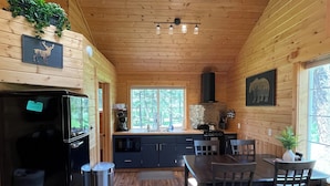 A wooden cabin interior with a kitchen, dining area, and a large window.