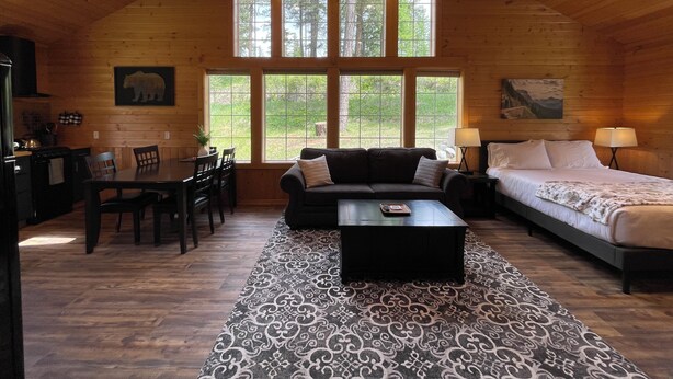 A cozy log cabin interior with a dining area, a sofa, a bed, and large windows offering a view of the outdoors.
