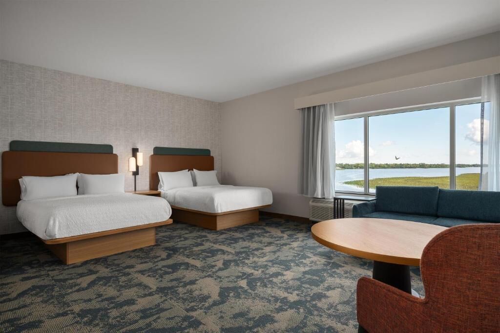 A hotel room with two beds, a sofa, a table, and a large window with a view of a body of water.