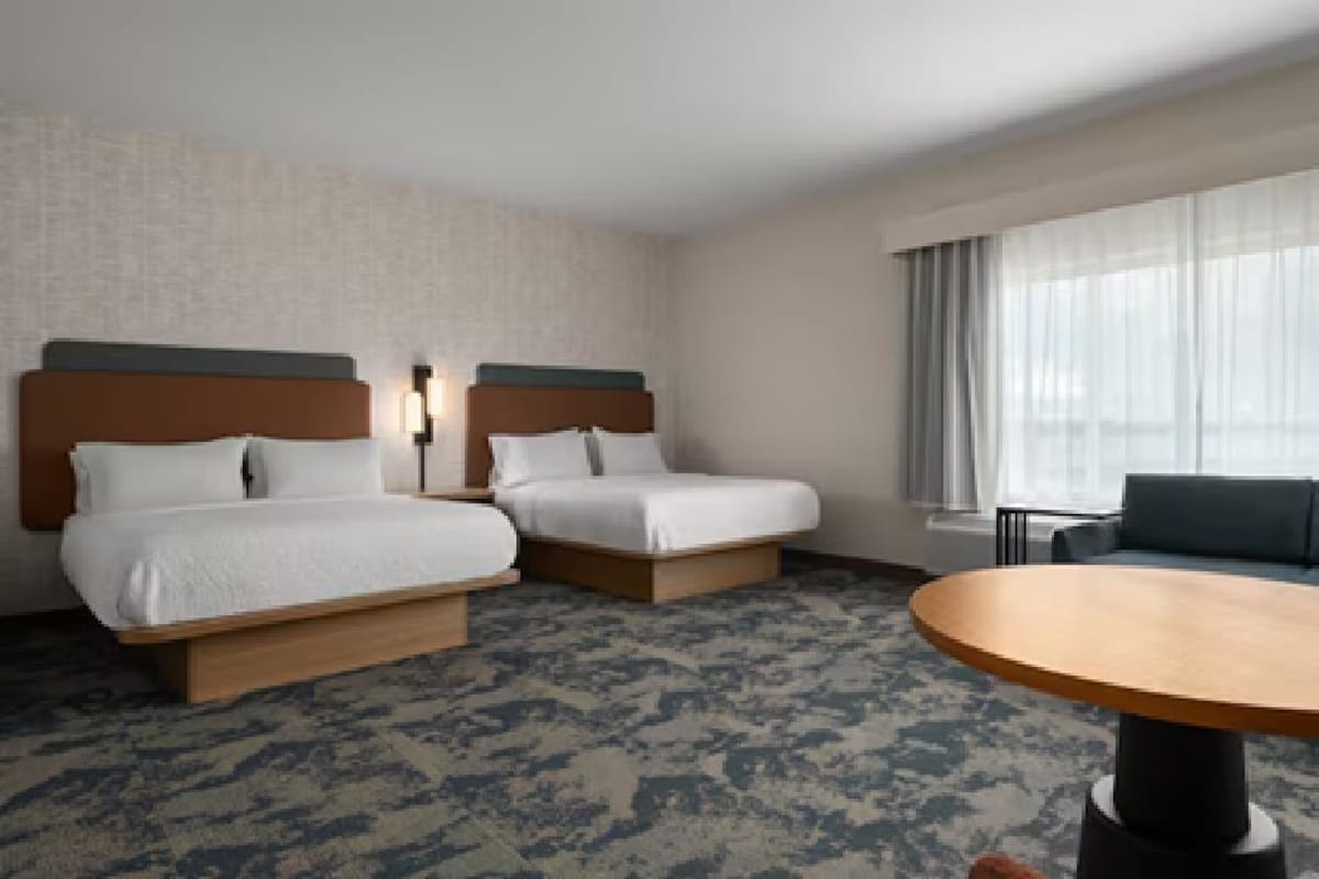 Ein Hotelzimmer mit zwei Betten, einer Schlafcouch, einem runden Tisch und einem großen Fenster mit Vorhängen.