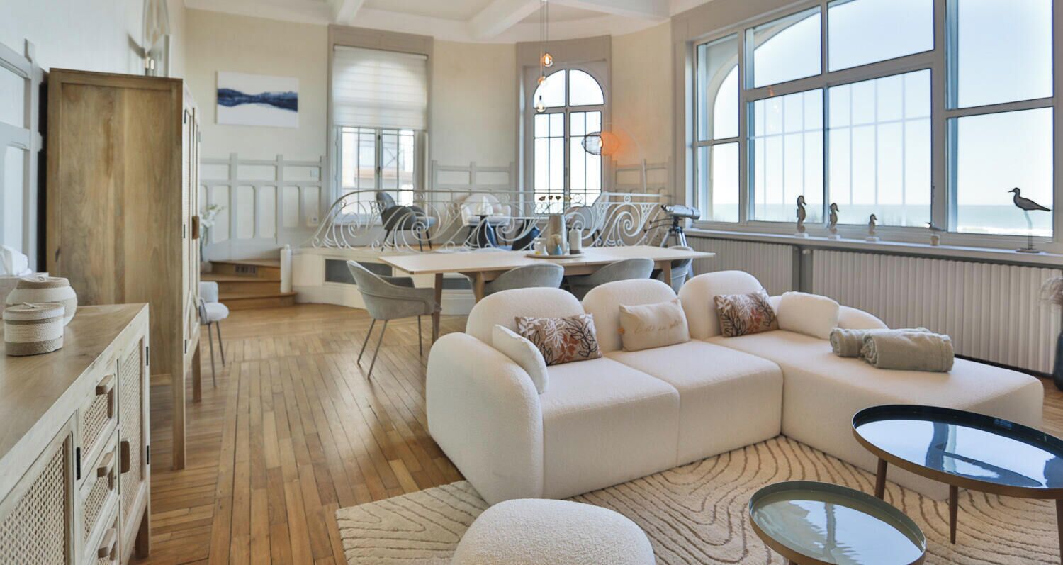 Un salon spacieux doté d’une grande fenêtre, un coin repas avec une table et des chaises, et un coin salon confortable avec un canapé et un pouf.