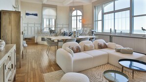 Een ruime woonkamer met een groot raam, een eethoek met tafel en stoelen, en een gezellige zithoek met bank en poef.