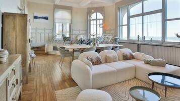 Un salon spacieux doté d’une grande fenêtre, un coin repas avec une table et des chaises, et un coin salon confortable avec un canapé et un pouf.