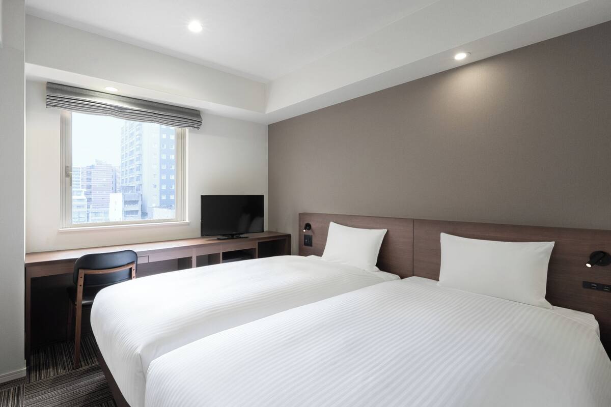 A hotel room with a large bed, a desk with a chair, a television, and a window with a view of buildings.