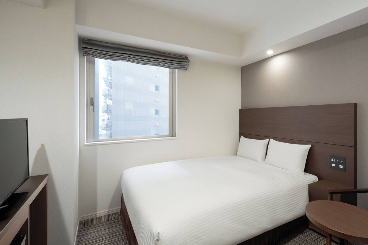 A hotel room with a bed, a TV, a chair, and a window with a view of buildings.