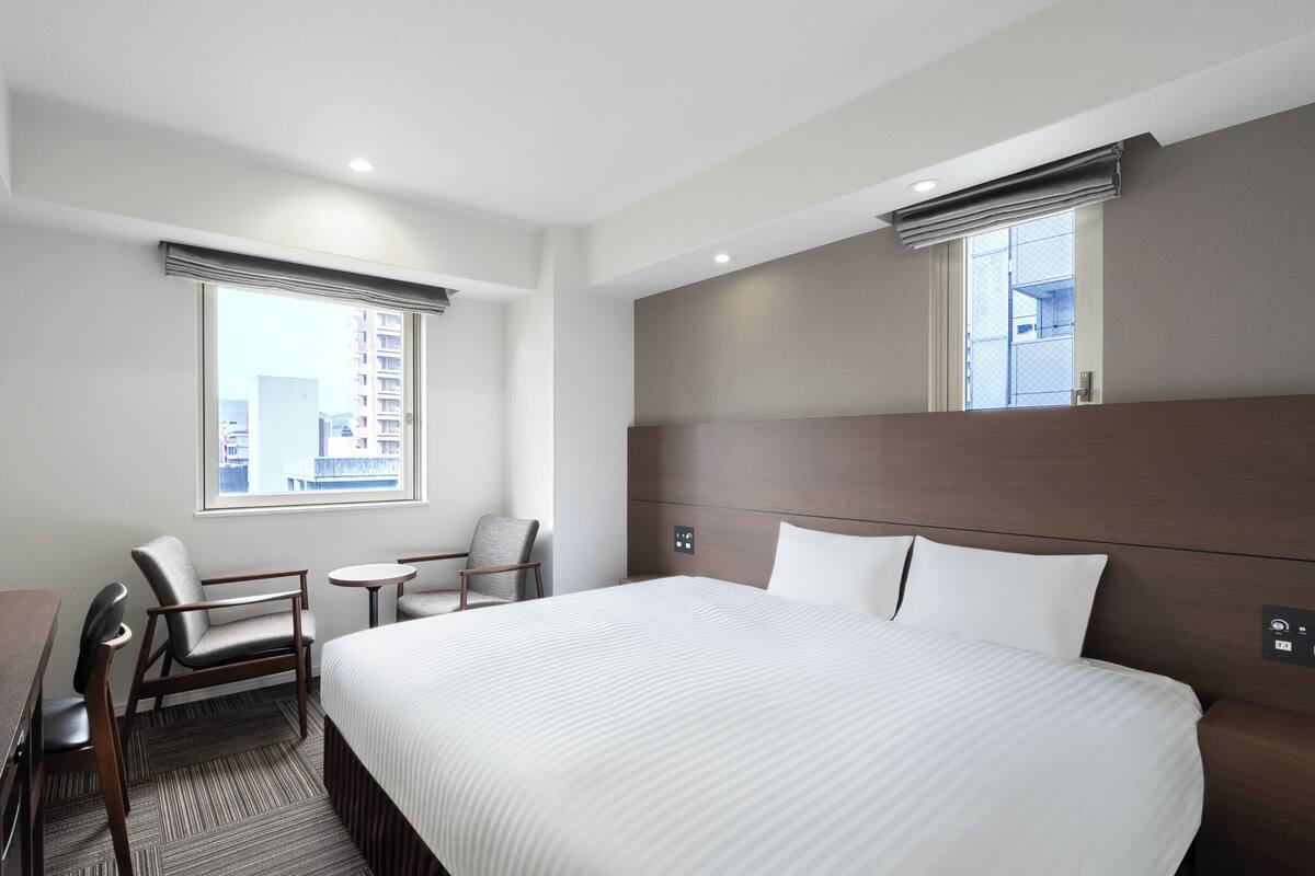 A hotel room with a large bed, a TV, a desk, two chairs, and a window with a city view.