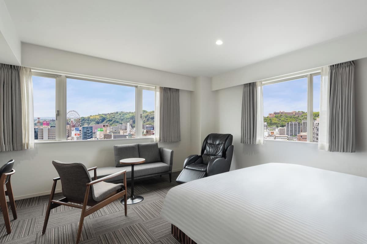 A hotel room with a bed, a desk, a chair, and a massage chair. There are two windows with curtains, and a view of a cityscape.