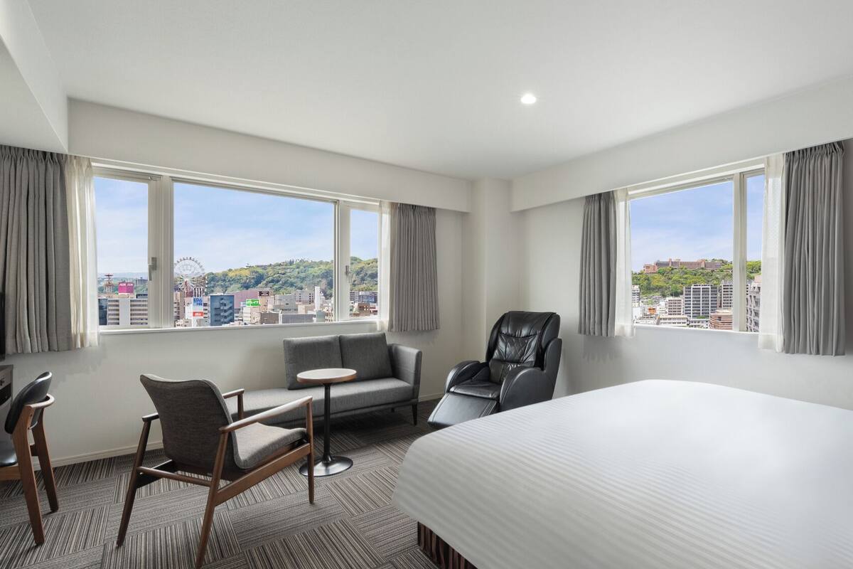 A hotel room with a bed, a desk, a chair, and two windows offering a city view.