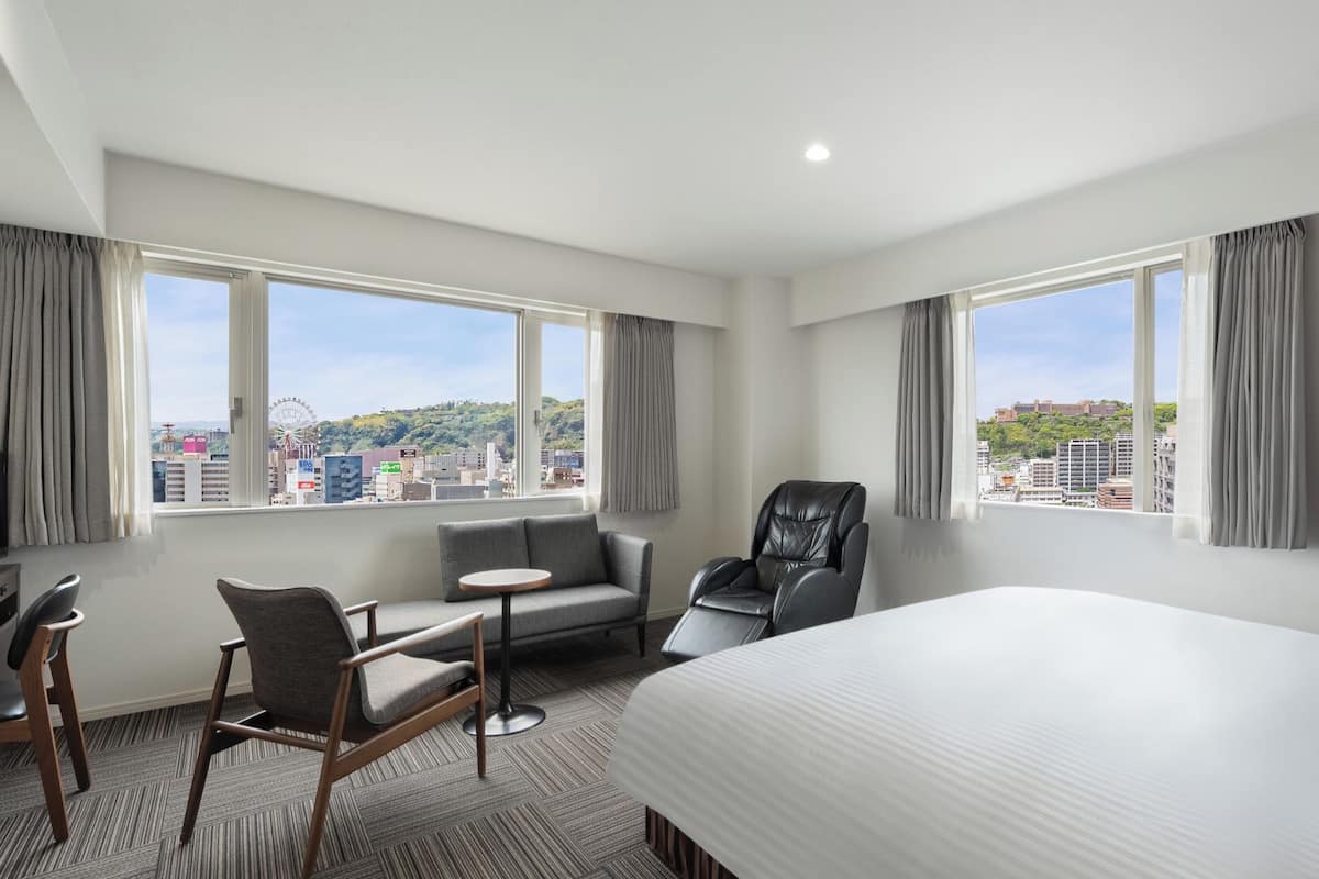 A hotel room with a bed, a desk, a chair, and two windows offering a city view.