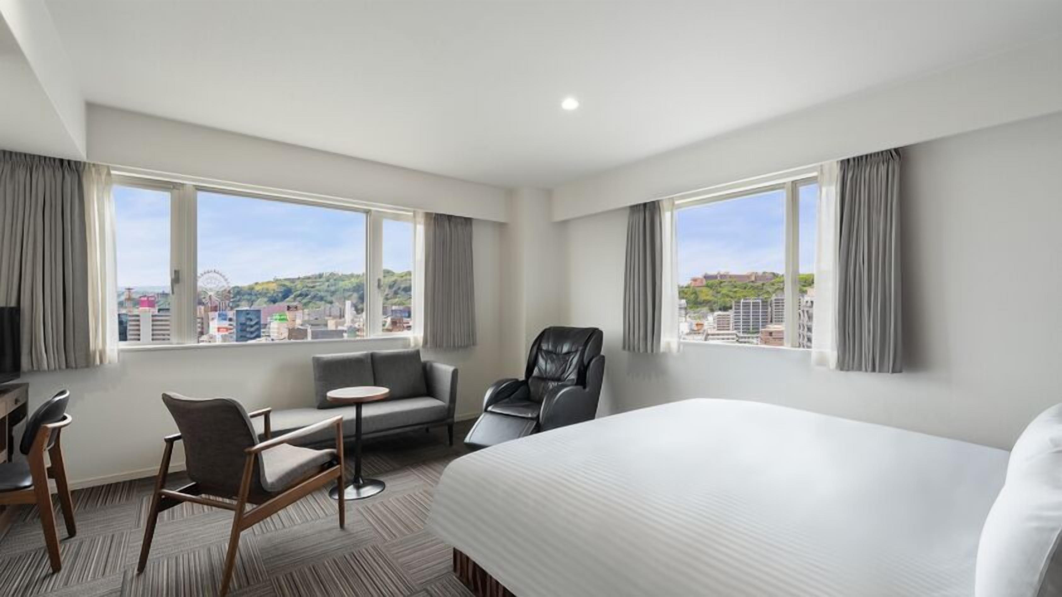 A hotel room with a bed, a desk, a chair, a sofa, and large windows offering a city view.