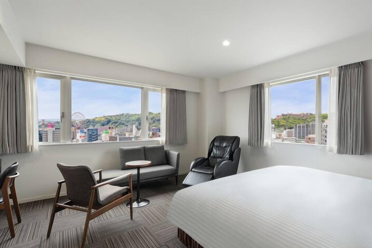 A hotel room with a bed, a desk, a chair, a sofa, and large windows offering a city view.