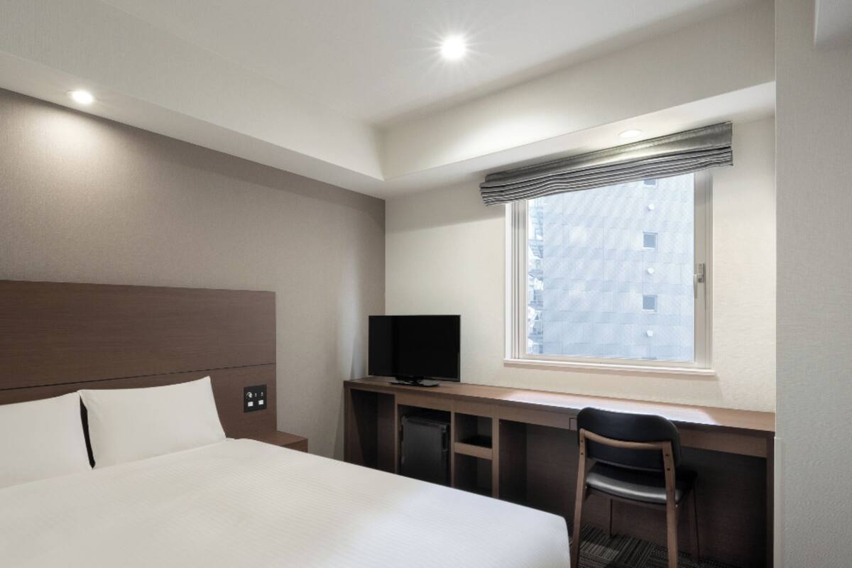 A hotel room with a bed, a desk with a TV, a chair, and a window with a view of buildings.