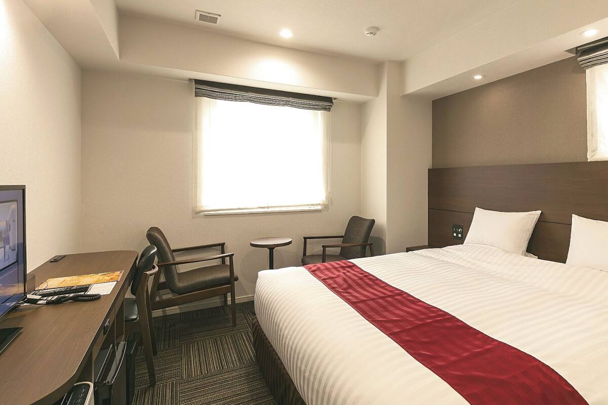 A hotel room with a bed, two chairs, a desk with a computer, and a window with curtains.