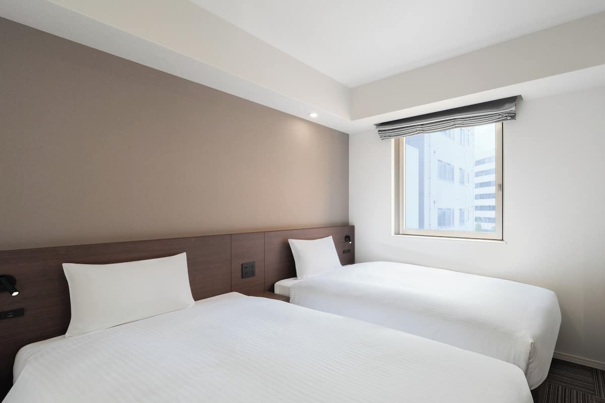 A hotel room with two beds, a television, and a window with a view of buildings.