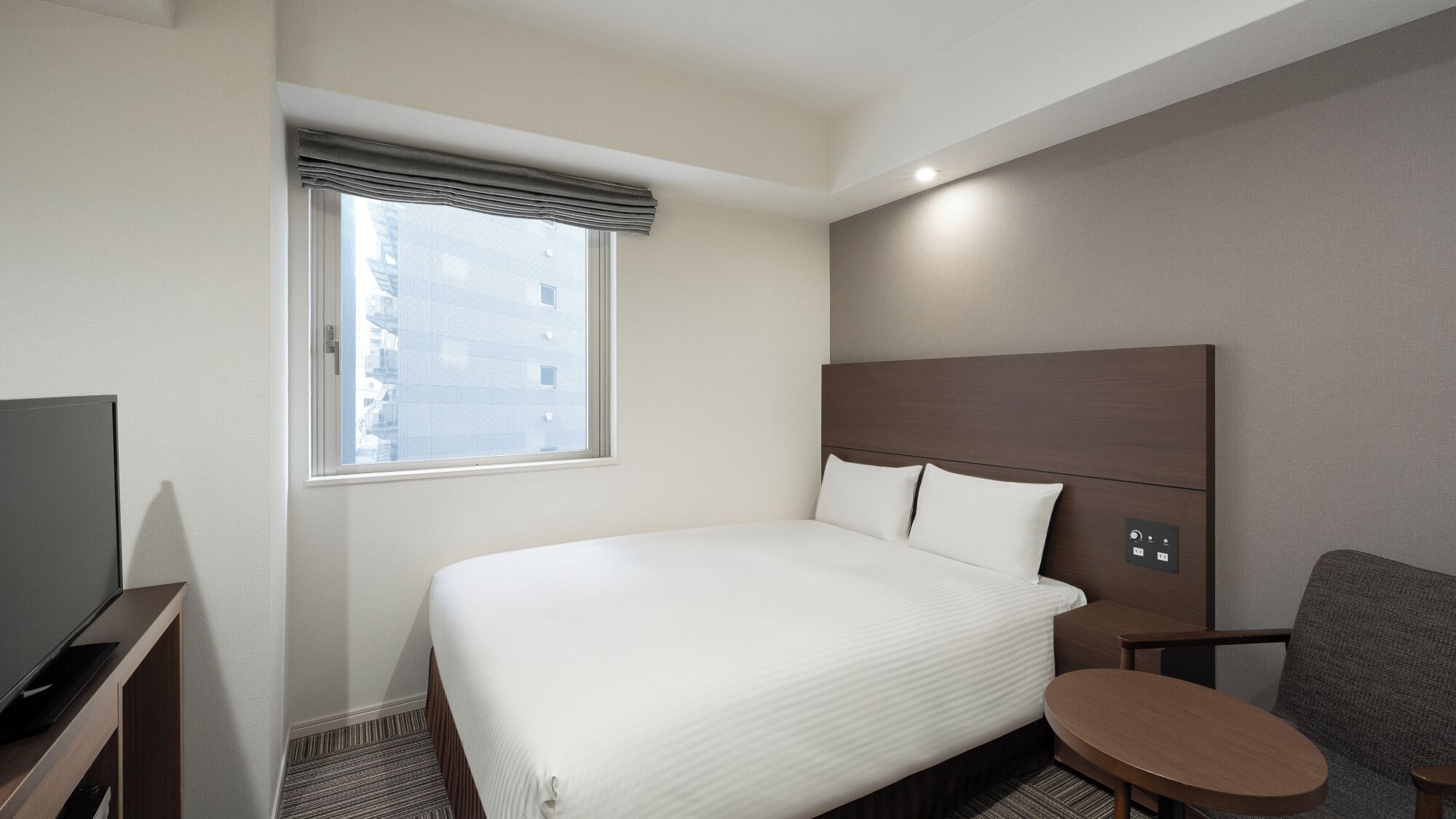 A hotel room with a large bed, a television, a small table, and a window with a view of buildings.
