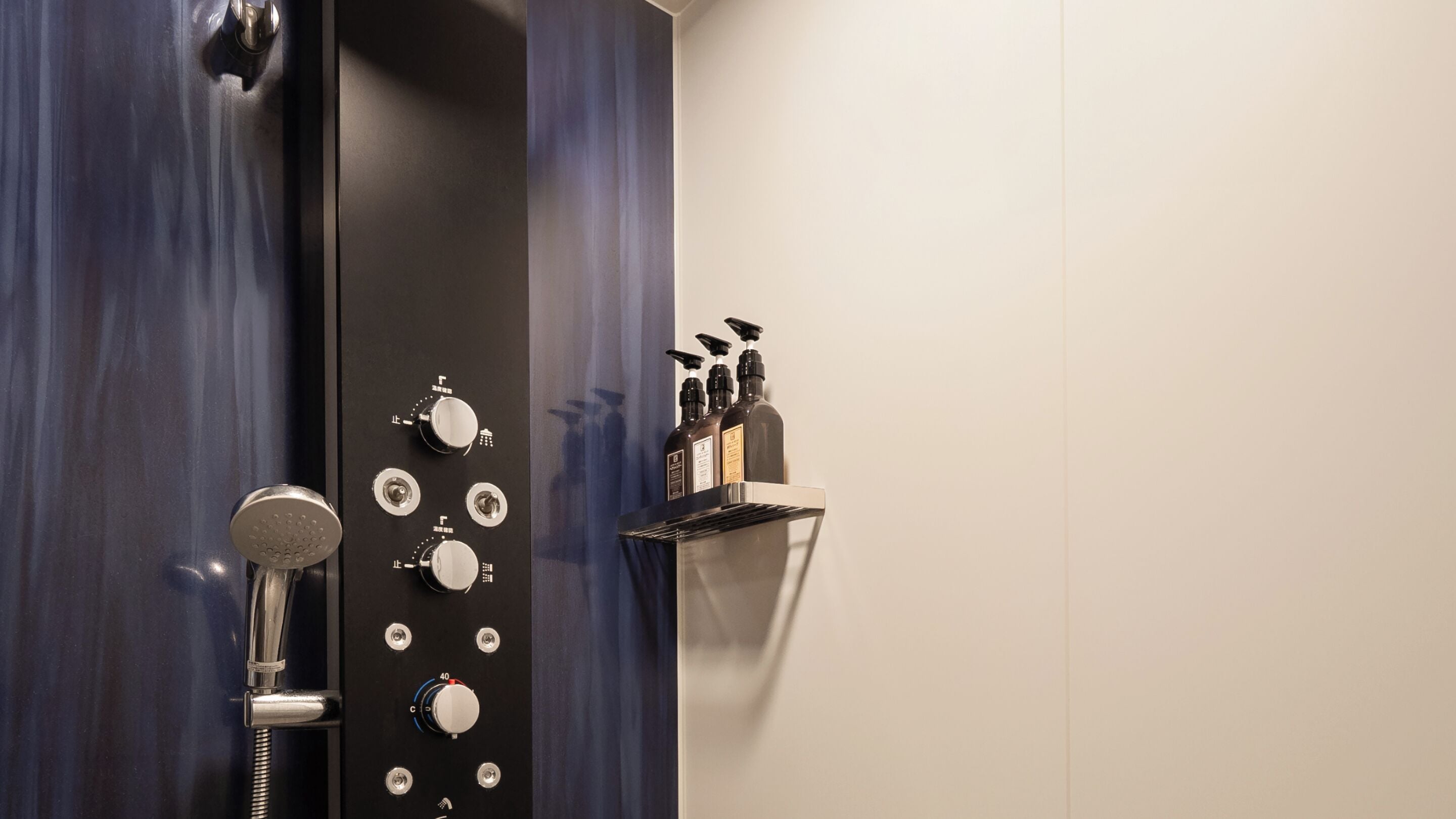 Un baño con panel de control de ducha, cabezal de ducha de mano y estante con artículos de aseo.