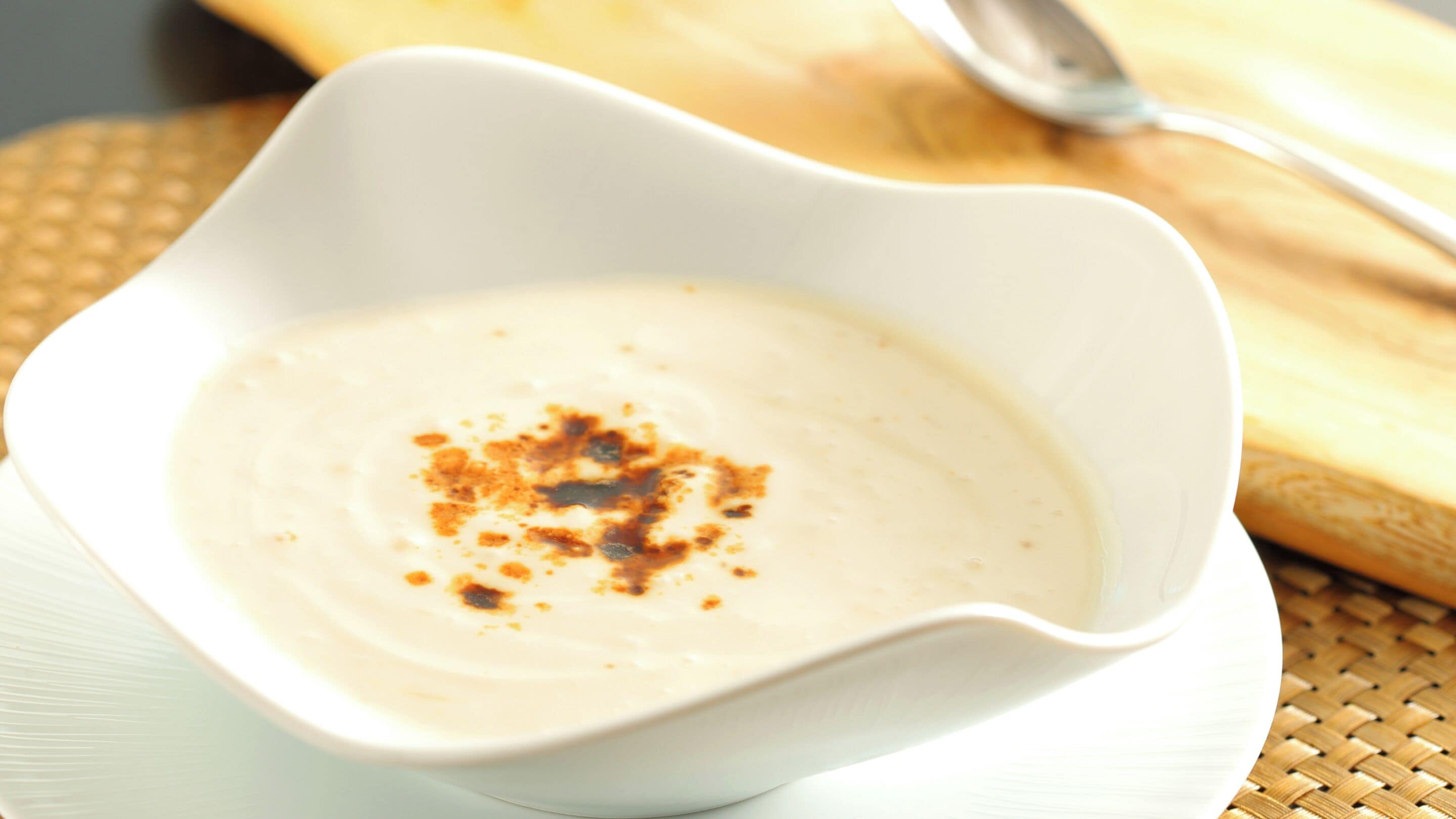 A white bowl of creamy soup with a sprinkle of spice on a woven mat.