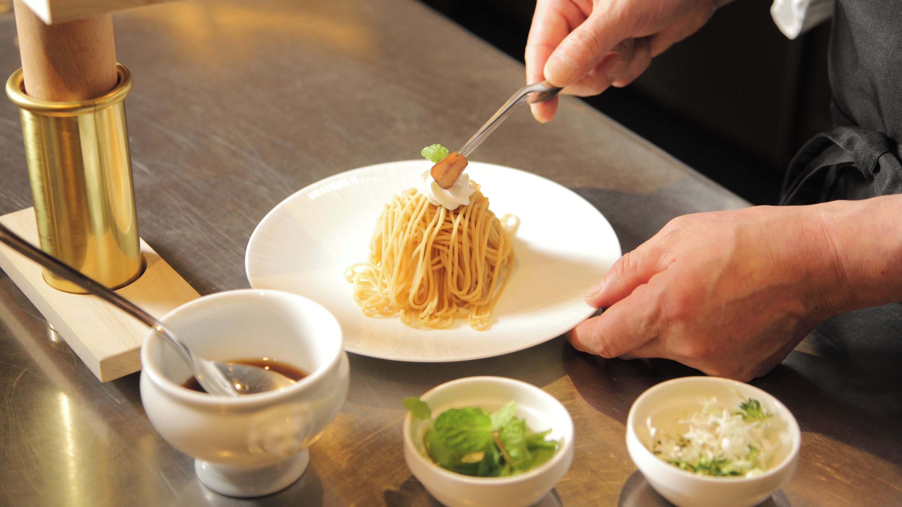 Une personne servant des spaghettis à l’aide d’une fourchette dans une assiette blanche, accompagnés de petits bols de sauce, de légumes verts et de fromage râpé.