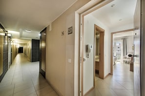 A hotel hallway with a modern design, featuring a tiled floor, a door with a phone, and a visible room number.