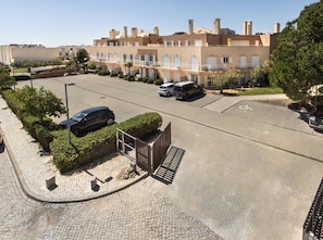 A parking area with a paved road, a black car, and a building with multiple windows and balconies in the background. - Paradise: Private Balcony with Pool View (Cabanas de tavira)