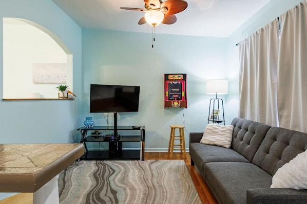A living room with a flat-screen TV, a ceiling fan, a small table with a plant, a sofa, and a patterned rug.