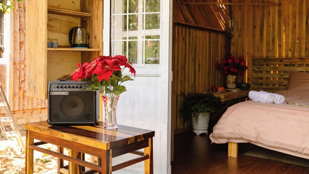 A wooden cabin interior with a bed, a table with a radio and flowers, and a window.