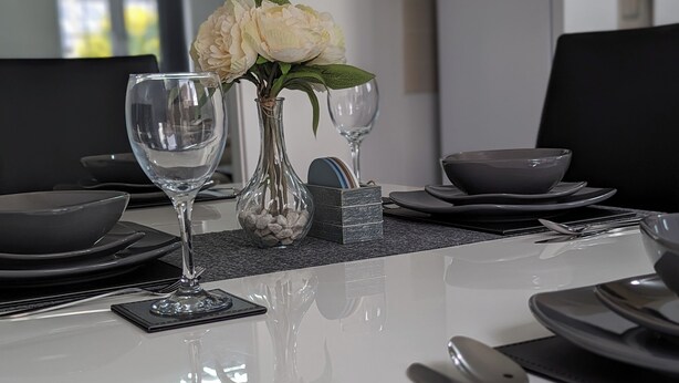 Ett modernt matbord med tillhörande stolar, dekorerat med genomskinliga glas, en vas med vita blommor samt svarta och grå tallrikar och skålar.