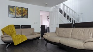Ett vardagsrum med två beige soffor, en gul pläd, en trappa och en klocka på väggen.