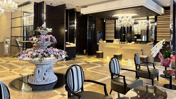 A modern hotel lobby with a reception desk, seating area, chandeliers, and a decorative floral arrangement. - Graces Hotels (Inzai)