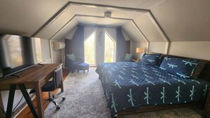 A bedroom with a sloped ceiling, a bed with a patterned bedspread, a desk with a chair, and a window with curtains. - Private Home in the White Mountains! (Campton)