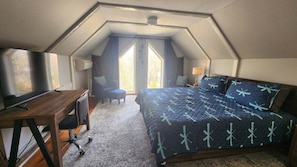 A bedroom with a sloped ceiling, a bed with a patterned bedspread, a desk with a chair, and a window with curtains.