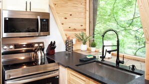 A modern kitchen with a stainless steel microwave, oven, and kettle, wooden cabinets, a black countertop, and a window with a view of greenery. - Modern A-Frame-Hot Tub-Gaming Area-EV Charger-10 Minutes to Slade (Stanton)