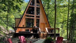 A wooden cabin with a triangular roof, surrounded by a gravel area with red chairs and a fire pit. - Modern A-Frame-Hot Tub-Gaming Area-EV Charger-10 Minutes to Slade (Stanton)