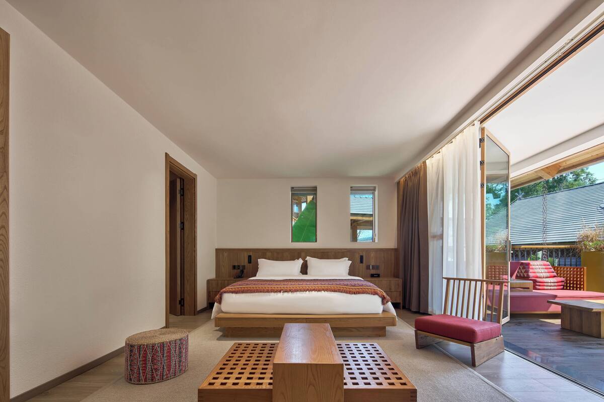A modern bedroom with a large bed, wooden headboard, bedside tables, a bench, and a view of the outdoors.