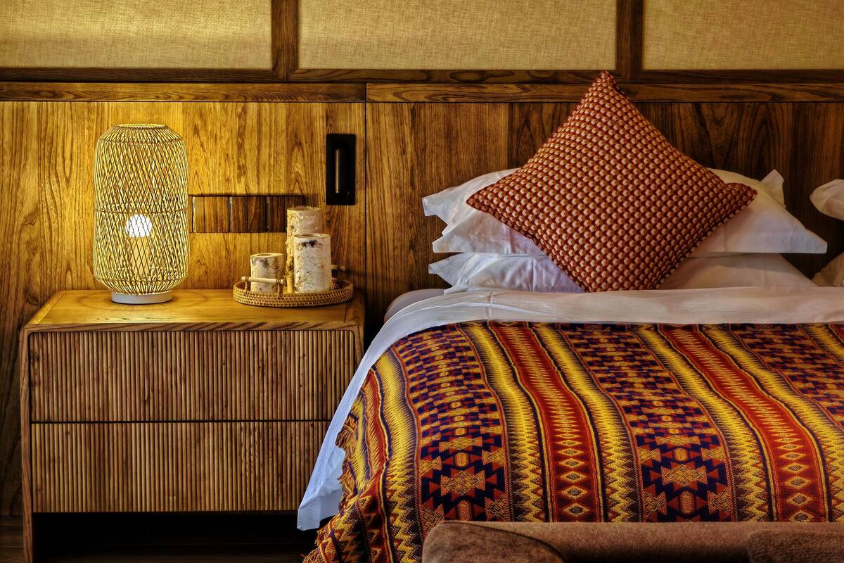 A wooden nightstand with a woven lamp, a patterned throw blanket, and a bed with pillows.