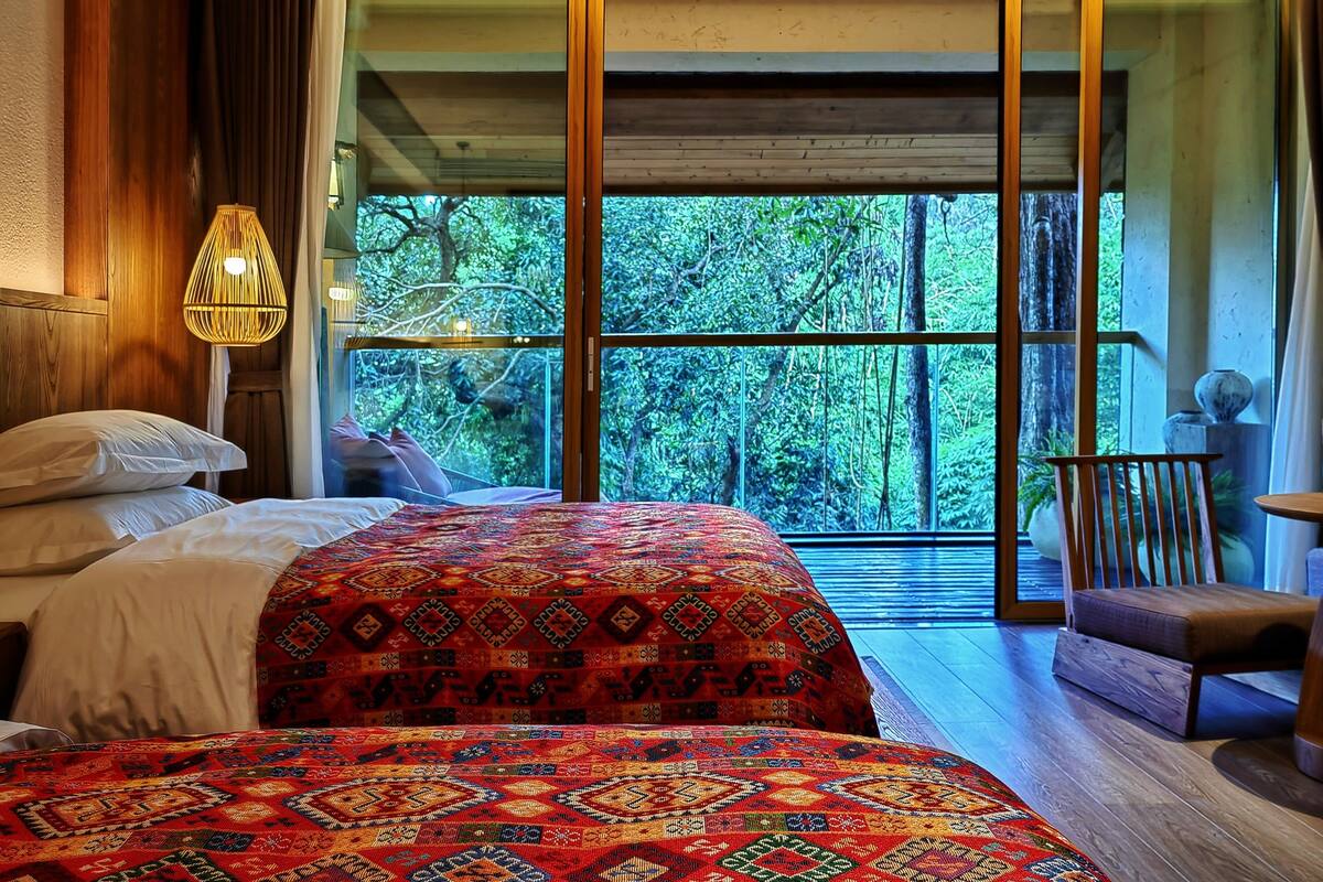 A bedroom with a large bed, a wooden headboard, a hanging light fixture, and a view of the outdoors through large windows.