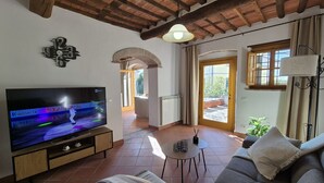 En stue med et fladskærms-tv, en sofa, et lille bord og en lampe. Der er et vindue med gardiner og udsigt til det fri.