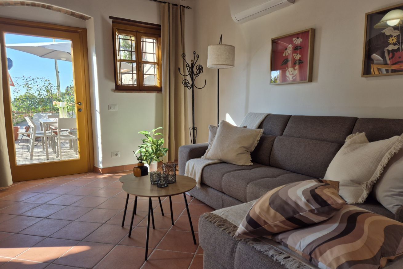 Una sala de estar con sofá, una mesa de centro, una lámpara de pie y una puerta que da a un área exterior con sillas y sombrillas.